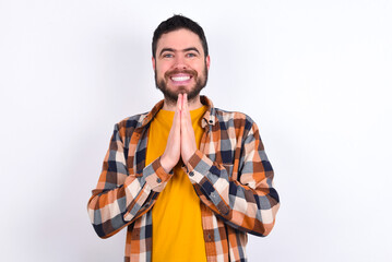 young caucasian man wearing plaid shirt over white background keeps palms together, has pleased...