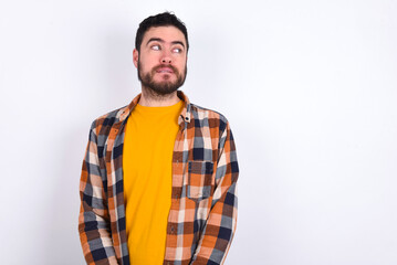 young caucasian man wearing plaid shirt over white background with thoughtful expression, looks away keeps hands down bitting his lip thinks about something pleasant.