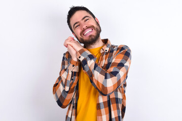 Dreamy young caucasian man wearing plaid shirt over white background with pleasant expression, closes eyes, keeps hands crossed near face, thinks about something pleasant