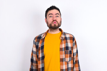 young caucasian man wearing plaid shirt over white background, keeps lips as going to kiss someone, has glad expression, grimace face. Standing indoors. Beauty concept.