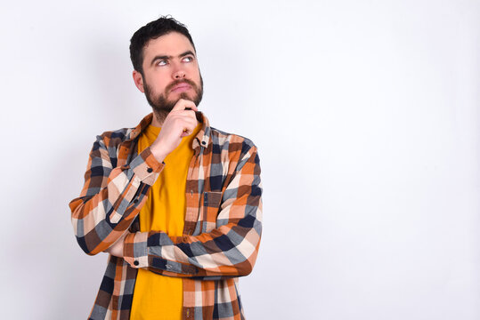 Shot of contemplative thoughtful young caucasian man wearing plaid shirt over white background keeps hand under chin, looks thoughtfully upwards.