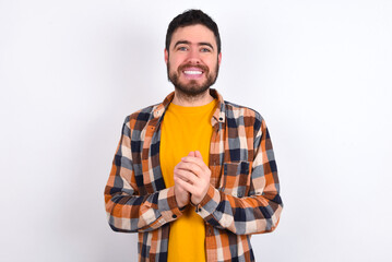 Happy young caucasian man wearing plaid shirt over white background stands against orange studio wall keeps hands on heart, swears be loyal, expresses gratitude. Honesty concept.