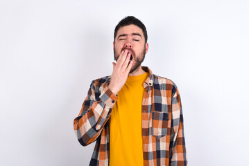 Sleepy young caucasian man wearing plaid shirt over white background yawning with messy hair, feeling tired after sleepless night, yawning, covering mouth with palm.