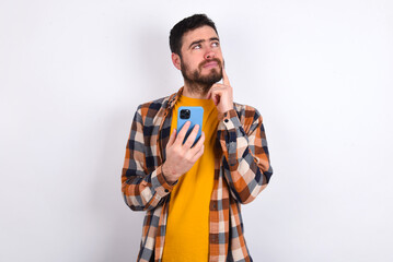 Thinking dreaming young caucasian man wearing plaid shirt over white background using mobile phone and holding hand on face. Taking decisions and social media concept.