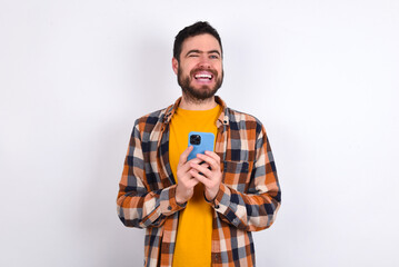 Pleased young caucasian man wearing plaid shirt over white background using self phone and looking and winking at the camera. Flirt and coquettish concept.