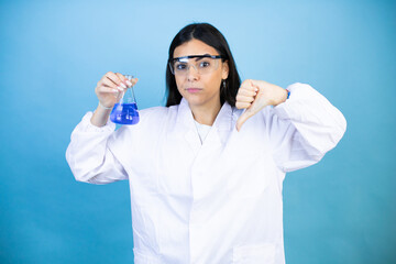 Young brunette woman wearing scientist uniform holding test tube over isolated blue background with angry face, negative sign showing dislike with thumb down