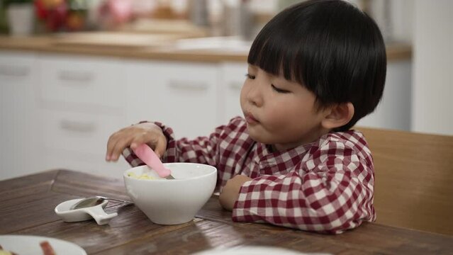 closeup of unhappy Asian little boy making a disgust expression while looking at green vegetable in bowl and shaking head show dislike at dining table