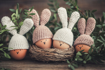 Four Easter eggs in crochet hats with rabbit ears in a nest on a wooden table. Soft focus.
