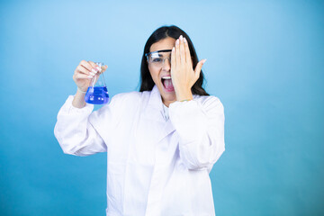 Young brunette woman wearing scientist uniform holding test tube over isolated blue background covering one eye with hand, confident smile on face and surprise emotion.