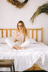 Happy smiling young attractive caucasian woman in eyeglasses using smartphone, sitting on a bed.