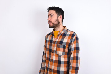 young caucasian man wearing plaid shirt over white background stares aside with wondered expression has speechless expression. Embarrassed model looks in surprise