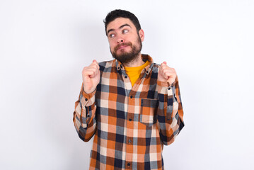 young caucasian man wearing plaid shirt over white background clenches fists and awaits for something nice happened looks away bites lips and waits announcement of results