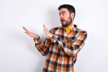 Displeased young caucasian man wearing plaid shirt over white background keeps hands towards empty space and asks not come closer sees something unpleasant