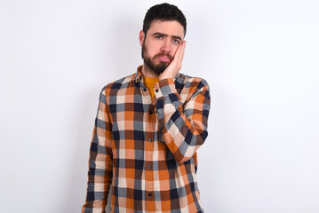 Sad lonely young caucasian man wearing plaid shirt over white background touches cheek with hand bites lower lip and gazes with displeasure. Bad emotions