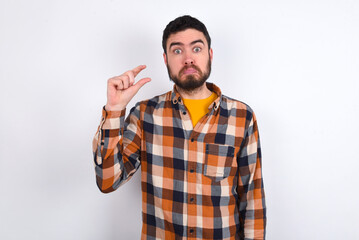 young caucasian man wearing plaid shirt over white background purses lip and gestures with hand, shows something very little.