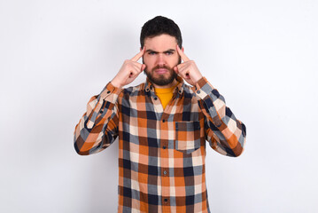 Serious concentrated young caucasian man wearing plaid shirt over white background keeps fingers on temples, tries to ease tension, gather with thoughts and remember important information for exam
