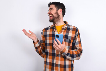 Happy pleased young caucasian man wearing plaid shirt over white background raises palm and holds cellphone uses high speed internet for text messaging or video calls