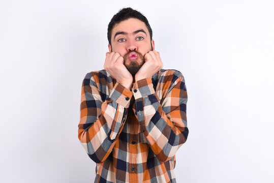 young caucasian man wearing plaid shirt over white background with surprised expression keeps hands under chin keeps lips folded makes funny grimace