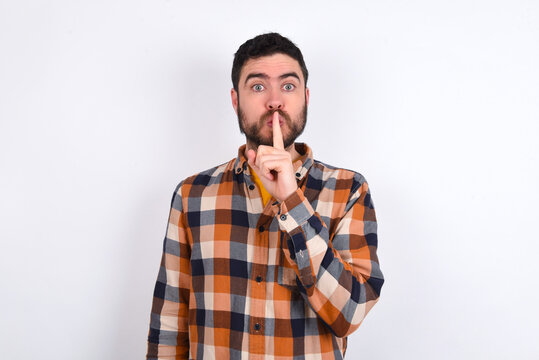 Surprised young caucasian man wearing plaid shirt over white background makes silence gesture, keeps finger over lips and looks mysterious at camera