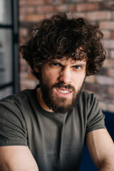 Close-up vertical headshot of angry handsome young man looking at camera sitting on chair in cozy living room with brick wall. Front view of confident curly male posing at home.