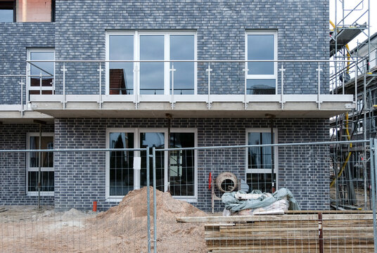Frontansicht: Detail einer Baustelle eines neu gebauten Mehrfamilienhauses hinter einem Bauzaun, mit Zementmischer, Material, Brettern, Sand, Kies, Werkzeug, Gerüst, selektiver Fokus, Copy Space