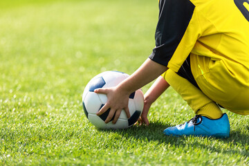 Kid pick up the ball from the playing field. Child holding soccer ball in hands. Boy in soccer uniform and cleats playing sports