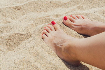 Female feet with red pedicure sunbathing on beach sand.