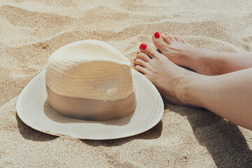 Hat and female feet with red pedicure on the beach sand.