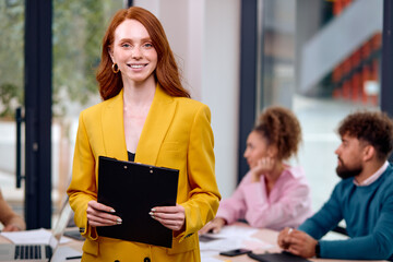Red-haired Smiling Woman With Clipboard In Hands Posing In Office During Business Meeting With...