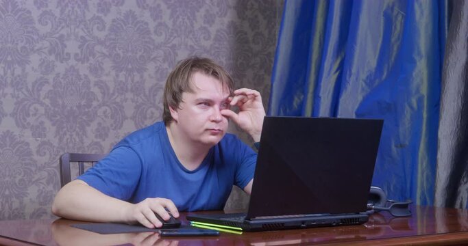 Young bored man with silly look sits at table, touches his eye and works or plays using laptop. Guy does not understand computers, so he broke something and opened his eyes wide in shock