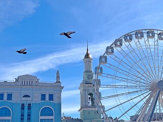 Kyiv city capital of Ukraine beautiful cityscape. Kiev old town peaceful scenic view.  Flying doves fly over the Ferris wheel. Clear sky at sunny day above Kontraktova square.