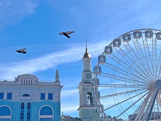 Wandcirkels plexiglas Kyiv city capital of Ukraine beautiful cityscape. Kiev old town peaceful scenic view.  Flying doves fly over the Ferris wheel. Clear sky at sunny day above Kontraktova square. © OLENA