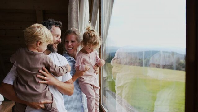 Happy young family in pajamas with small children looking at view from window on holiday.