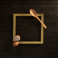 Egg shells, a wooden spoon and a gold frame on a dark wooden background. Text space. Top view. Minimal style.