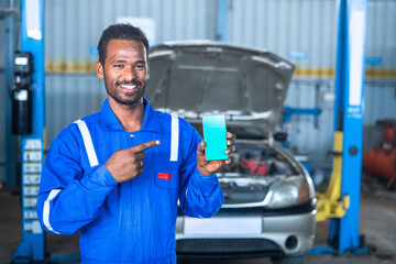 Smiling car mechanic showing green screen mobile phone with uniform in front of car by looking at...