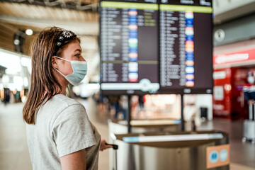Fototapeta na wymiar A young girl in mask waiting at the airport with visible flight information board