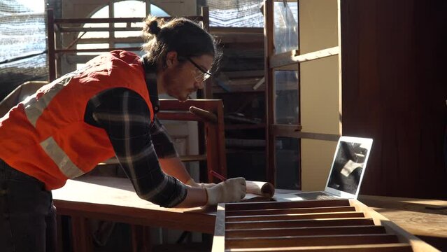 young hipster carpenter man working with computer laptop checklist in furniture workshop . craftsman wearing safety glasses checking order of clients or learning online