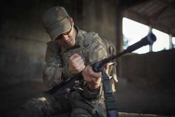 Fototapeta premium a Ukrainian soldier in a gray camouflage uniform wearing a cap and tactical black goggles sits on a concrete floor inside a large destroyed building and reloads a machine gun