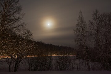 Front car lights on a snowy winter road in the countryside. Silhouette of forest with moon in cloudy sky, Sipoo, Finland.