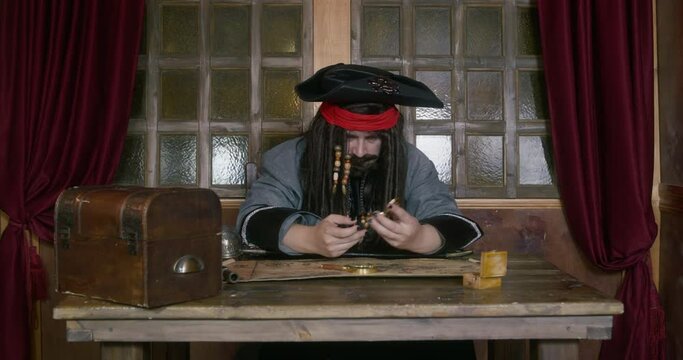 Pirate captain in cocked hat with long hair takes gold coin from treasure chest looking at map ON table against window in ship cabin
