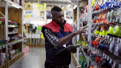 Male supervisor walking between rows in hardware store examining goods on shelves with tablet
