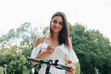 Fototapeta premium Pretty young mixed race brunette woman renting an electric scooter using mobile app outdoors. Smiling lady in white summer dress paying by transport using smartphone and looking at camera