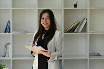 A portrait of a young smiley pretty Asian woman standing in the office looking to the camera with confidence  for business, finance and work concept.