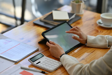 Cropped view young woman sitting in the office using a blank white screen tablet and  and a calculator working on data, charts and documents on the table, for business, finance and technology concept.