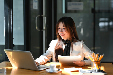 A portrait of a young pretty Asian woman sitting in the office using a laptop, calculator, working on the documents, for business, design, finance and technology concept.