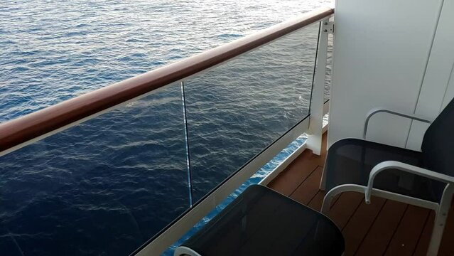 View of the open sea from a cruise terrace on a mid level deck above the life boats. Sea is calm. Terrace delimited by a wooden veranda has an empty chair and a small footrest.