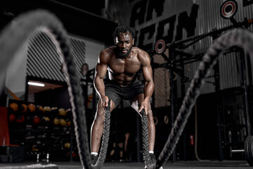 Portrait of strong young black man exercising with battle ropes during workout in modern gym,...