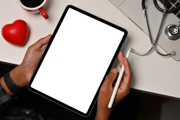Top view of hands holding a blank white screen tablet and a pen leaning on the white table with a heart model, laptop and a stethoscope on the sides for medical, health care and technology concept.