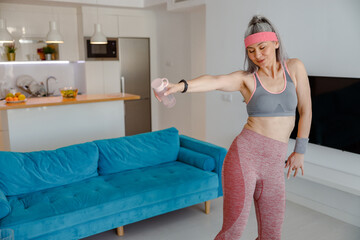 Smiling lady in sportswear holding bottle of water and dancing while doing exercise at home