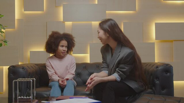 Caring positive pretty Asian female psychologist talking with upset adorable preadolescent African girl, helping to overcome mental problems and improve social skills during psychotherapy session.